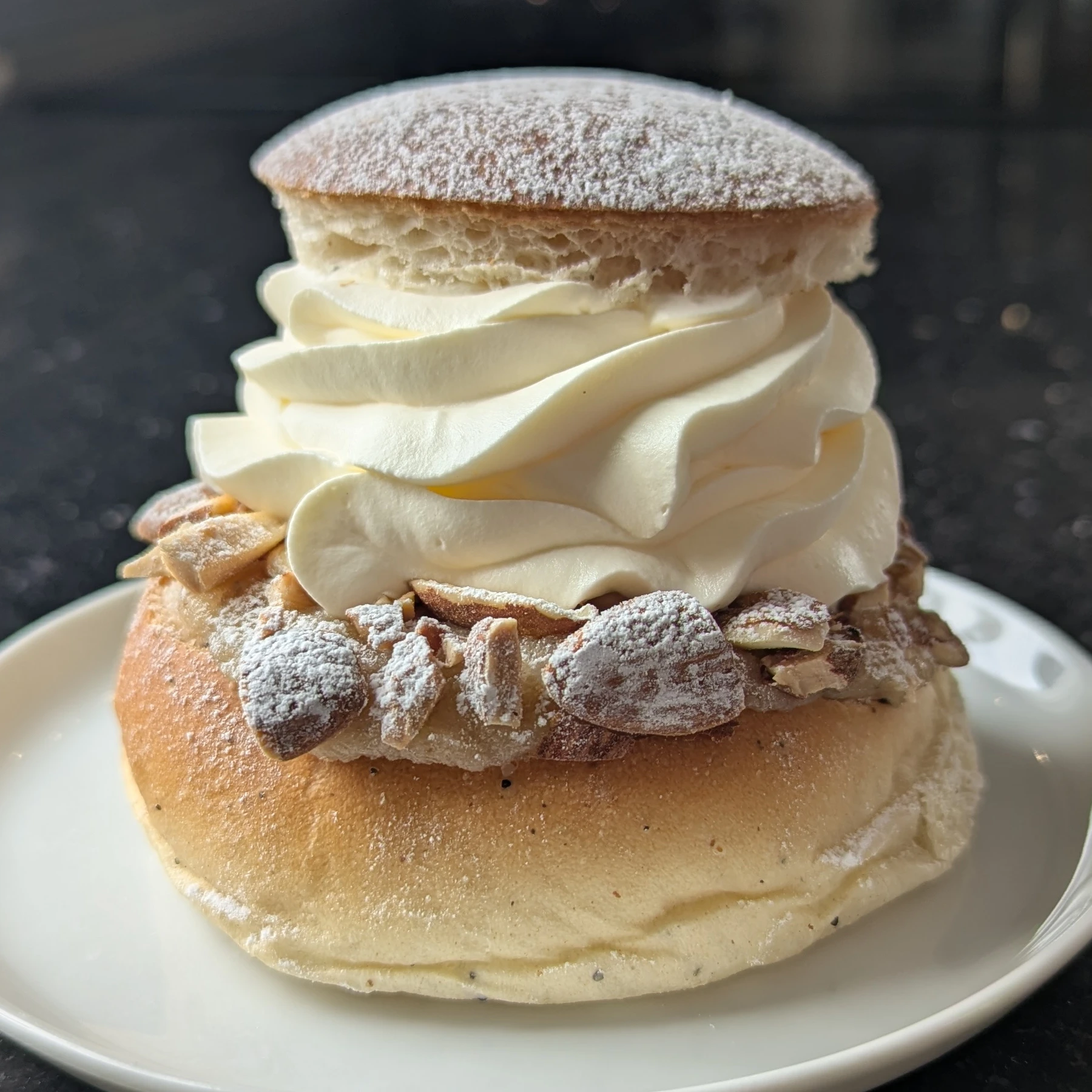 A semlor placed on a white round plate on a black stone surface. There are a lot of icing-sugar dusted roughly chopped almonds visible stuck to the almond paste. On top is a large swirl of very smooth cream piped in a anti-clockwise swirl using an open star piping tip. A round icing-sugar covered lid is perched on top. The bottom of the bun is slightly wrinkled and dots of cardamom can be seen. 