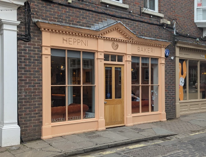The outside of Heppni Bakeri. It has a wooden front painted in a pale orange colour. There are lots of woodwork details on the pillars and at the top as well as muntins dividing up the windows into smaller sections. The words Heppni Bakeri are painted above the window along with an illustration of a croissant. In one of the windows is an illustration of a girl juggling four pastry products. 