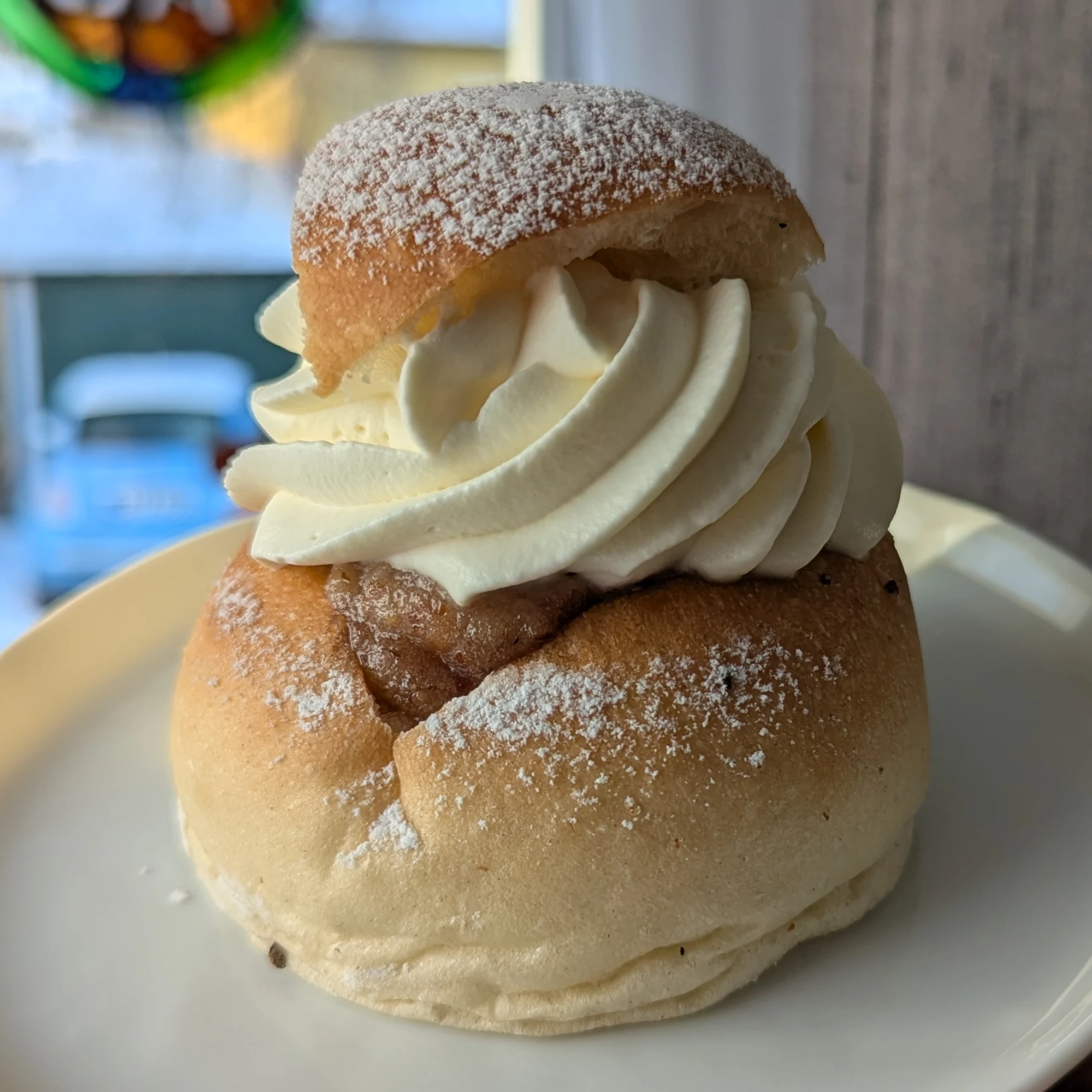 A semla with a triangular lid. The lid is dusted with icing sugar and placed so that the visible point is aligned with the cut in the bun where it came from. In the cut light brown almond paste can be seen. Some of the icing sugar has dusted the sides of the bun. The cream is a defined swirl, going clockwise from the lid. The fresh bun has wrinkles at the base as it has collapsed slightly from the weight of the filling.