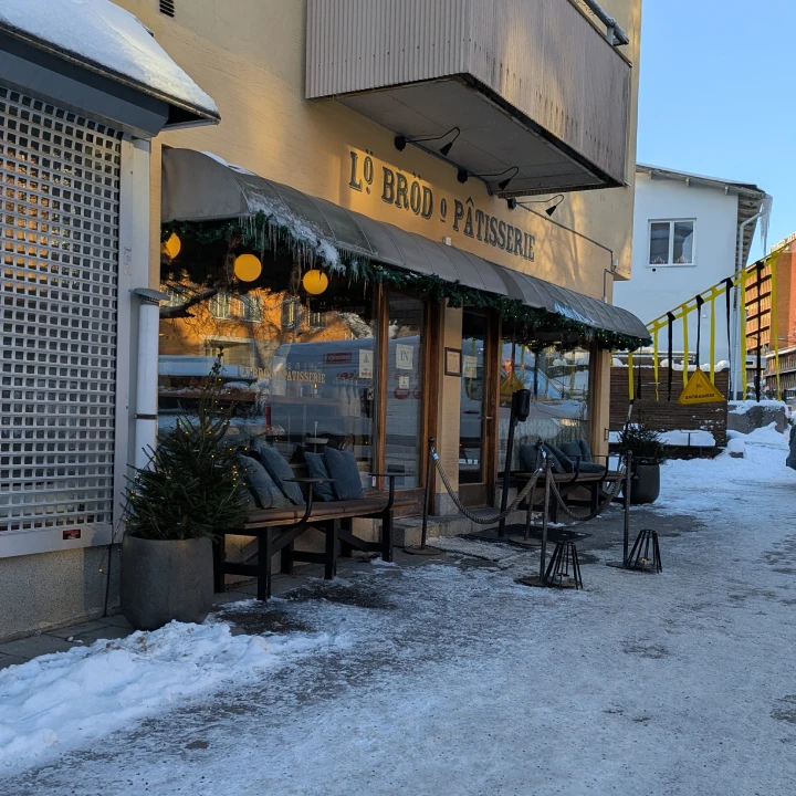 A view of the outside of Liningö bröd bakery. There is snow on the ground. Icicles can be seen hanging from the canopy. There is a Christmas tree to the right and one to the left and benches with cusions. Leading up to the entrance there is a rope railing on either side. At the very front are two candles in metal lanterns.