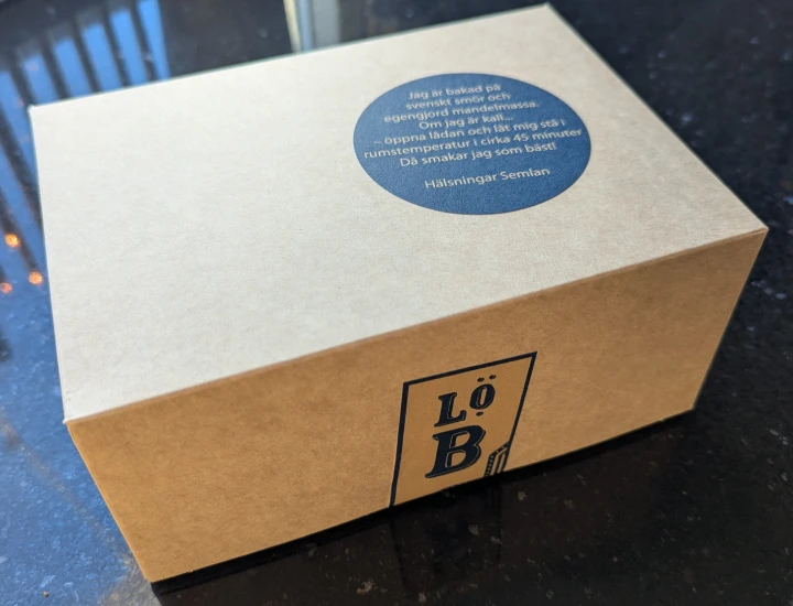 A brown cardboard box. The box is closed and sitting on a black marble kitchen surface. There are two stickers. The sticker on the top says (in Swedish) - "I am baked with Swedish butter and self-produced almond paste. If I am cold, open the box and let me stand at room temperature for about 45 minutes. Then I'll taste my best! Greetings, The Semla." On the front there is another sticker with the Lidingö bröd logo which is holding the box shut.