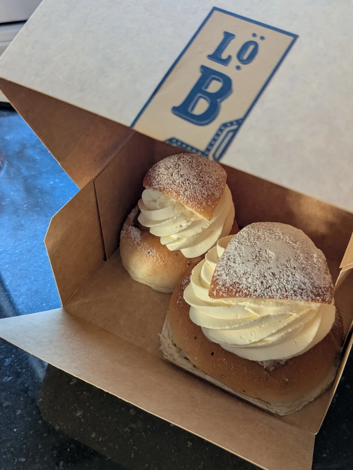 An open brown cardboard semla box. A Lidingö bröd sticker can be seen on the front of the lid. Inside the box are two semlor. One small towards the back left and one large towards the front right. The icing sugar on the top of the large semla is smudged where it has been touching the inside of the lid.