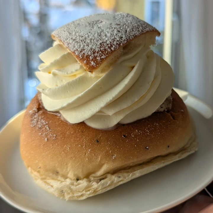A large semla with a triangular lid. The lid is dusted with icing sugar and placed so that the visible point is aligned with the cut in the bun where it came from. In the cut light brown almond paste can be seen. Some of the icing sugar has dusted the sides of the bun. The cream is a defined swirl, going clockwise from the lid. The cream is starting to melt slightly. The bun has visible spots of crushed cardamom and has wrinkles at the base as it has collapsed slightly from the weight of the filling.