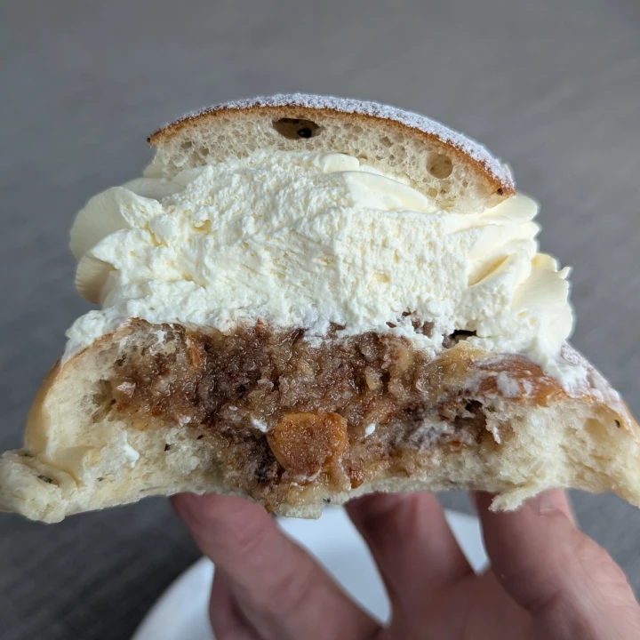 View of the inside of the semla. The cream is as high as the bun, with the lid on top. The almond paste is thick and  brown with large pieces of almond visible. One piece in the centre looks like it is a whole almond. Bits of cardamom can be seen in the bun. The semla is being held up by finger tops.