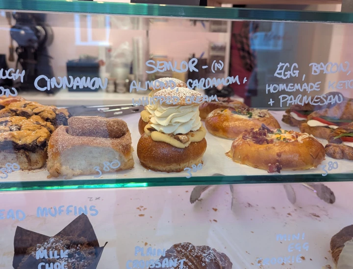 A glass counter inside the bakery. Behind the glass a number of bakery products can be seen. There is white writing on the glass giving the name of each product and the price. In the centre of the photo is the row of semlor. There are two semlor left.