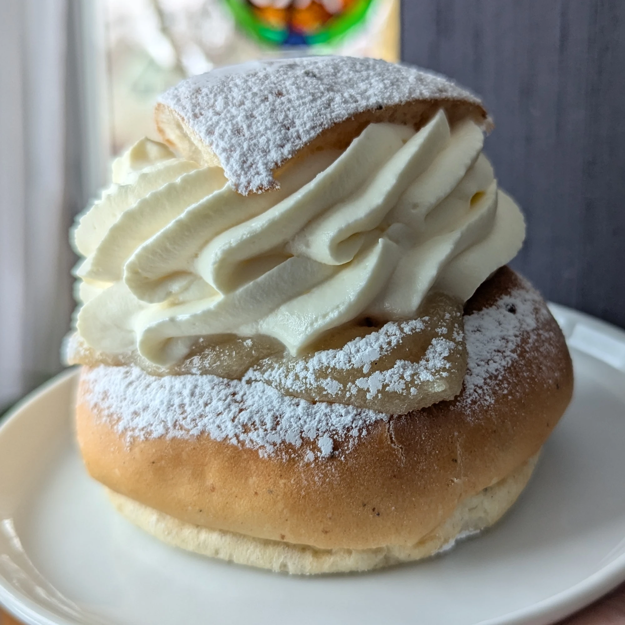 A semla with a heavily dusted triangular lid. Under the lid is a a swirl of cream that has been piped in two layers with lots of definition. Light coloured almond paste filling can be seen under the cream and along with the bun has also received a dusting of icing sugar. The semla is sat on a white round plate and is being held up in front of a window and has been photographed in natural light.