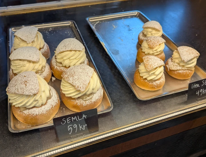 Two trays of semlor behind the glass counter. The left tray has 5 large semlor and a sign saying Semla 59kr. The right tray has 4 semlor and a sign saying small semla 59kr.