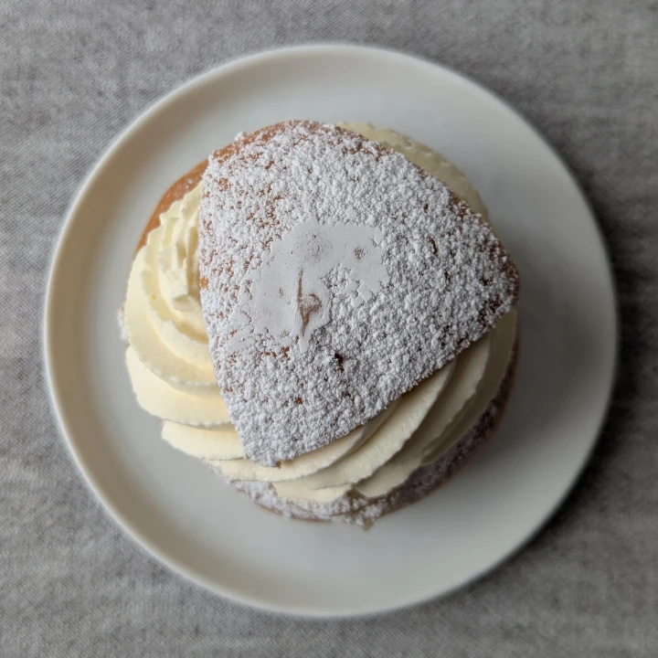 A view of the semla from above. There is a triangular lid that has been heavily dusted with icing sugar. The icing on the very top of the lid has been smudged where it has touched the top of the inside of the lid of the box it was transported in. Under the lid an excellently piped swirl of cream can be seen. The semla sits on a white round plate on a table surface with a grey table-cloth.