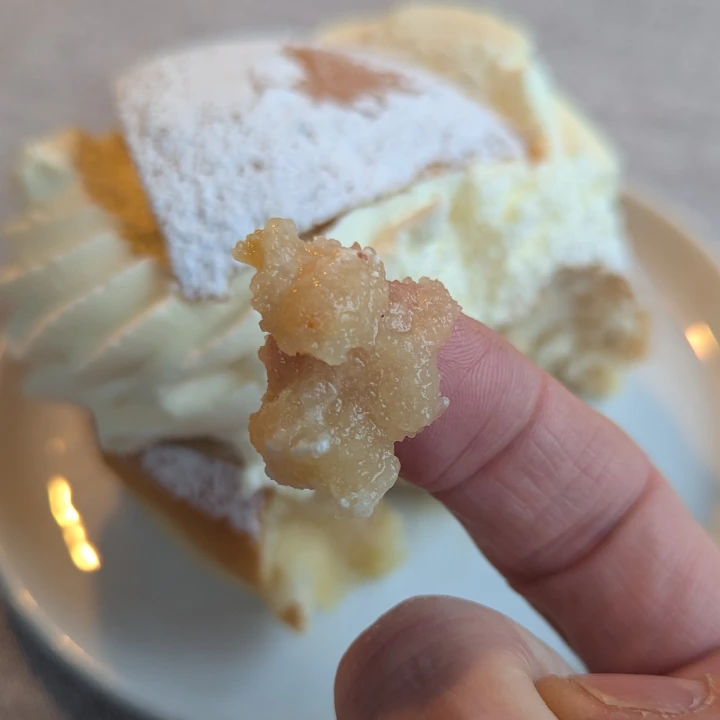 A finger with almond paste on it is held up towards the camera. In the background the semla can be seen. The paste on the fingertip is lightly colour and some lumps can be seen although the lumps are the same colour as the rest of the paste.