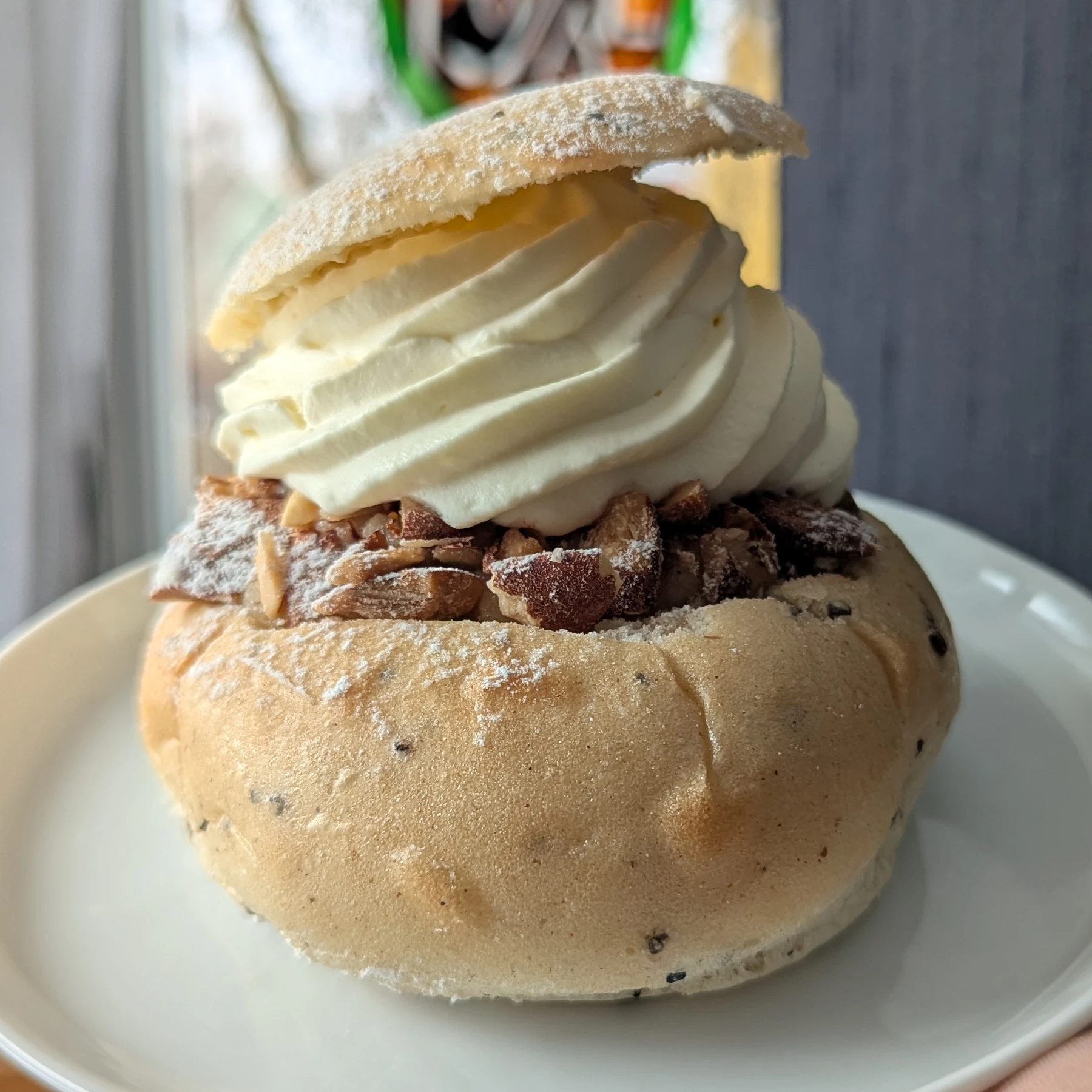 A pale semla. There are lots of black dots of cardamom, some crushed, some whole kernals. The lid is sitting at an angle on top of a swirl of piped whipped cream. There are lots of almonds visible, so many that the almond paste can't really be seen. There is a dusting of icing sugar on the lid, nuts and the top edges of the bun. The bun is sitting on a white plate. 