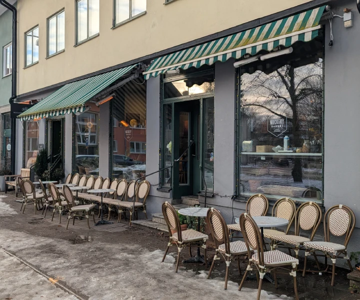 A view of the outside of Nockeby Bageri. Above the large shop windows are green and white striped awnings, one of which is extended. Ice and slush covers the pavement. There are round tables outside with rows of chairs that have wicker backs and seats.