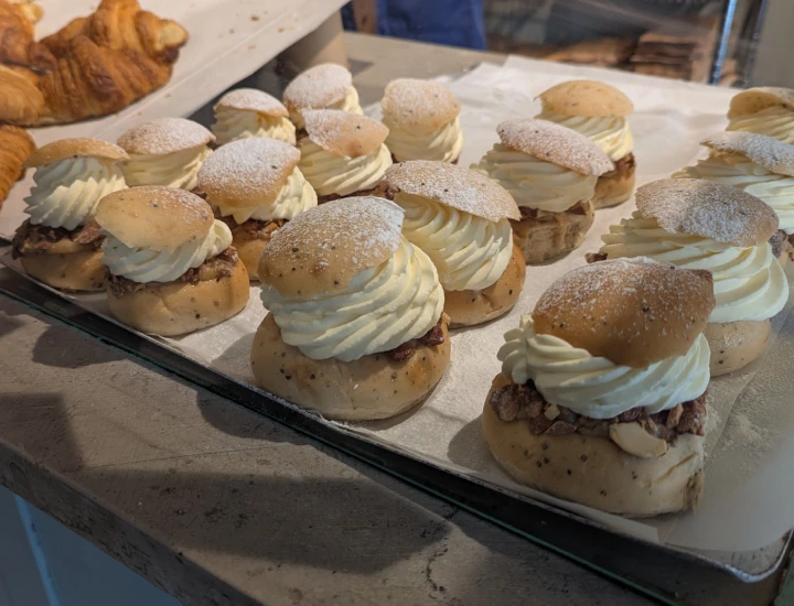 A tray of semlor on the counter of the bakery. To the left of the tray are 8 small semlor. To the right are 8 large semlor. The semlor are not identical in appearence. Lids are roughly cut and sitting at different angles. Some of the semlor have almost no icing sugar dusted on top. To the far left, a couple of coissants from the next tray along on the counter can be seen.