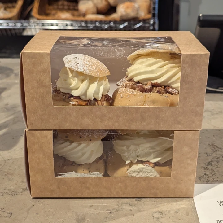Two semlor boxes with plastic windows stacked on top of each other on the counter of the bakery. The boxes are of brown cardboard and have plastic windows. The semlor can be seen inside. In the bottom box, the semla to the right has been squished against the plastic window leaving a smear of cream.