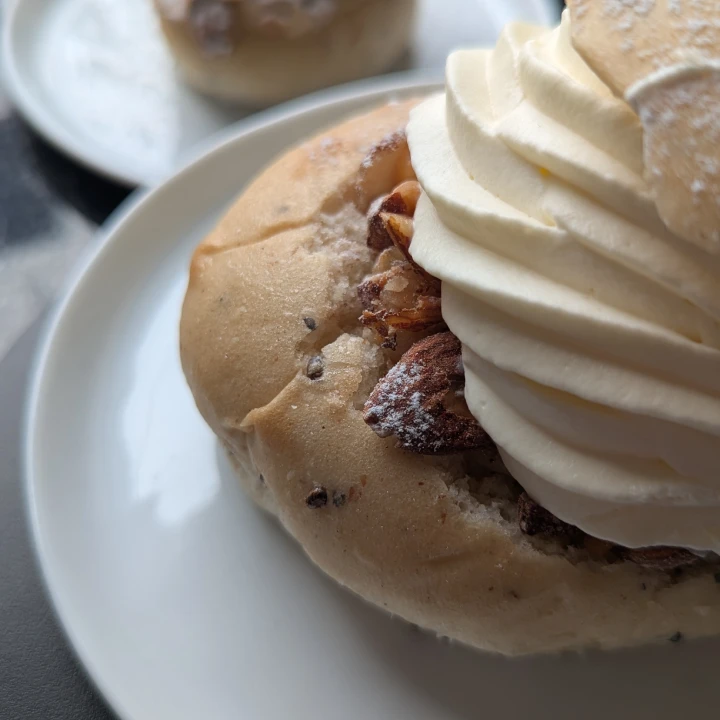 A close up of the large semla. The semla is untidily constructed with the bun. The well cut into the bun can be seen without paste. The whipped cream has been piped off-centre. The lid is sitting on the side of the cream. Large bits of cardamom can be seen on the pale semla bun.