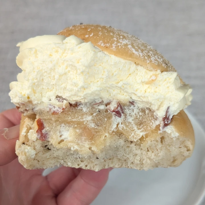 A view of the inside of a semla. The almond paste is pale and beige. Roughly chopped nuts can be seen on top of the paste. There is a large amount of cream, which is almost as high as the bun. The semla is being held by fingertips.