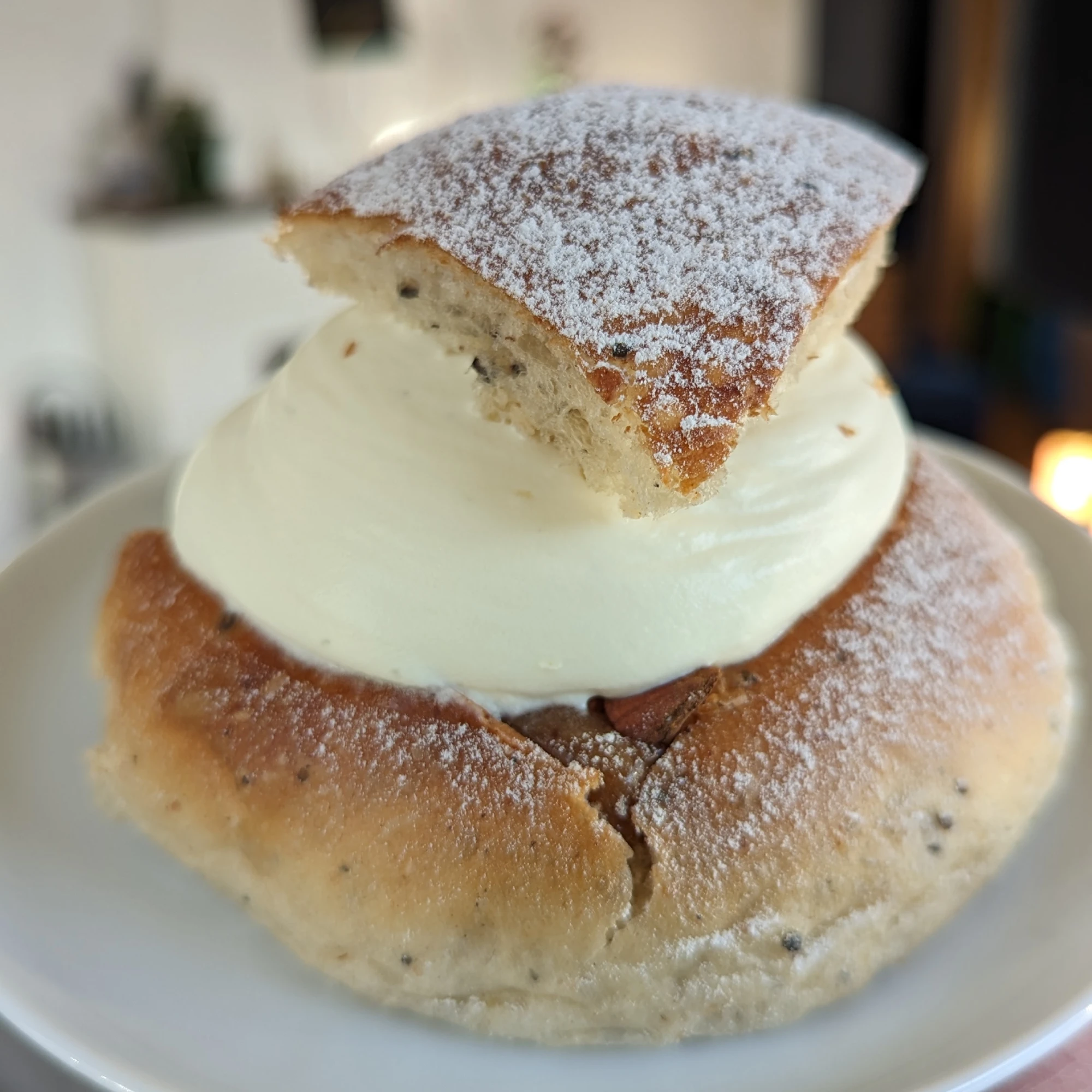 A semla with slightly wholemeal looking bun, smooth cream, and a triangular icing sugar dusted lid. There is a crack at the front of the semla where the well for the paste has been cut out. Inside the crack, the dark brown almond paste can be seen as well as a chopped almond.