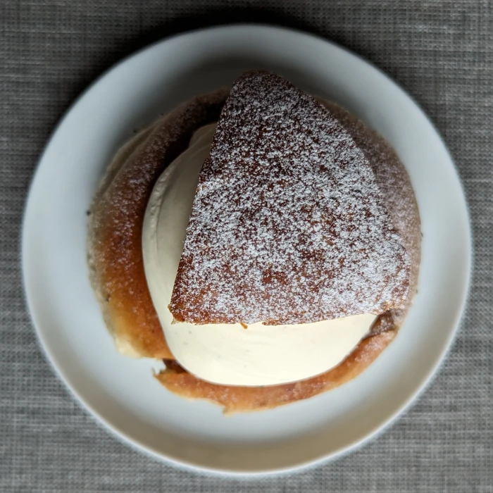 View of the semla from above. The triangular lid dusted with icing sugar sits on top of a smooth mound of whipped cream. The semla sits on a white circular plate, which is on a grey table cloth.