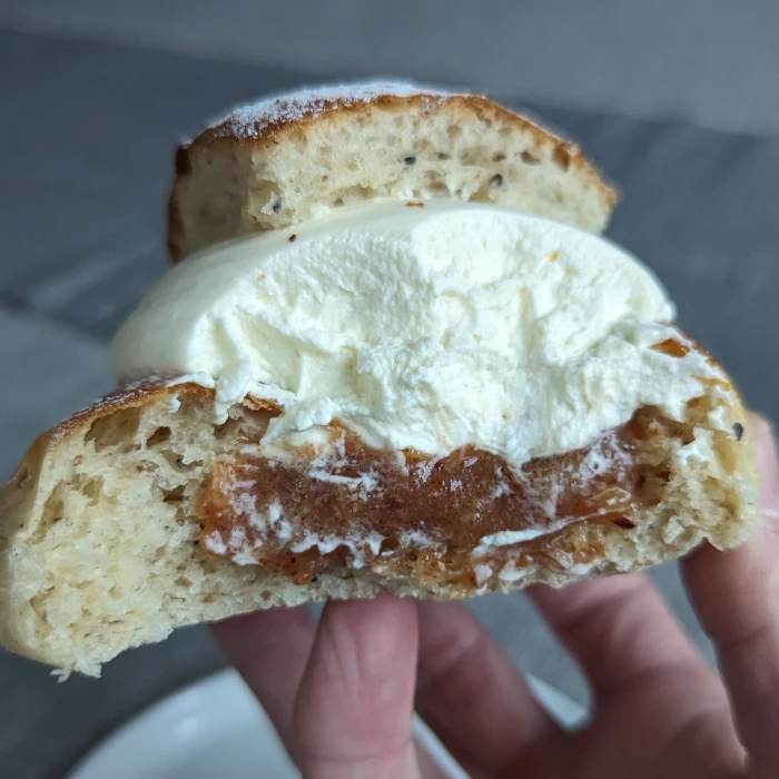 View of the inside of the semla.  The almond paste is thick and dark brown. Bits of cardamom can be seen in the bun. The semla is being held up by finger tops.